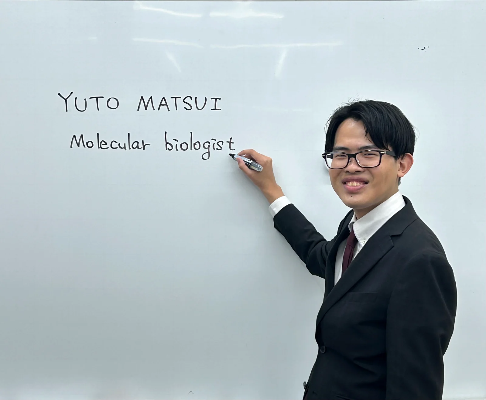 ホワイトボードに研究分野を書き示すスーツ姿のYuto Matsui